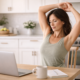Woman with dark hair sitting at a kitchen table stretching her arms overhead in natural light, representing gentle movement and core support